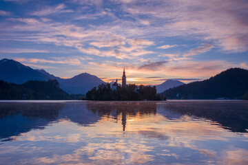 Beautiful sunrise and reflection at Lake Bled with the lake island and charming little church dedicated to the Assumption of Mary, famous tourist attraction in Slovenia - obrazy, fototapety, plakaty