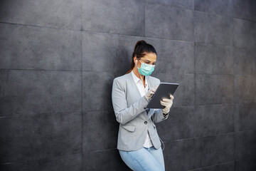 Portrait of cute businesswoman with protective mask and gloves on leaning on the wall and using tablet. Protection form corona virus concept.