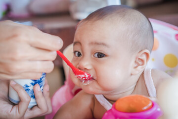 Cheerful cute baby eating baby food on a baby chair. Mother feeding food her baby girl with a red spoon. Happy Asian family at home and living lifestyle indoors. Healthy nutrition for kids concept