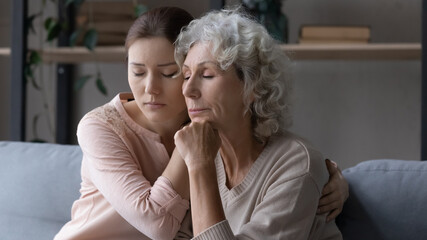 Unhappy senior Caucasian mother and grownup daughter sit on sofa hugging make peace reconcile,...