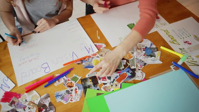 Group of women sitting at table and making vision board collages together to visualize their goals, wishes and plans for future during female training workshop, top view panning shot