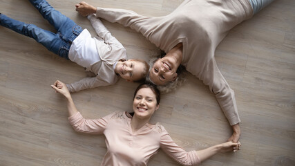 Top view portrait of overjoyed three generations of women lying on warm floor holding hands showing family unity and support, smiling loving girl with young mom and senior grandmother relax at home