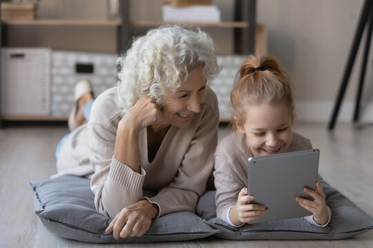 Smiling Mature Grandmother Lying On Floor With Little Granddaughter Using Modern Tablet Gadget Together, Happy Senior 60s Grandparent Relax Have Fun With Small Grandchild, Watch Video On Pad At Home