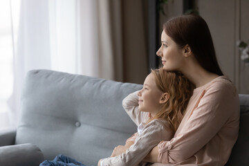 Fototapeta premium Young Caucasian mother and small preschooler daughter relax on sofa at home look in distance dreaming together, dreamy mom and little girl child hug cuddle, dream or visualize on couch in living room