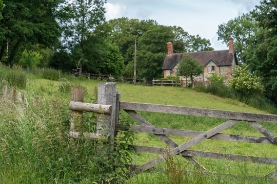 A Gate Into A Field In The English Countryside With A Stone Cottage In The Background.
