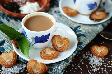 A cup of hot tea with cookies are placed on a piece of cloth. Cookies and sugar are spread all over the wooden table. The earthen plate contains the ingredients. This elevates the mood of tea.