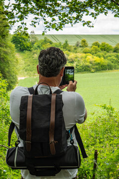 A man hiking in the English countryside stops at a beautiful serene view point to take a photo with his smart phone.  Image of man from behind taking a picture with his cell phone.