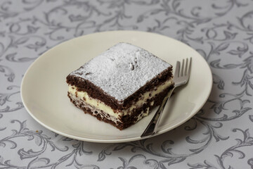 Dessert in a square shape interlaced with whipped cream and decorated with sugar. The dessert is on a white plate with a desert fork.