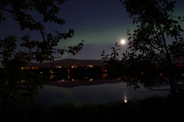 Naklejka premium majestic aurora borealis, northern light over calm mirror lake at night with buildings in the background
