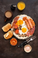 English traditional breakfast on a dark brown rustic background. Top view, flat lay. Fried eggs with bacon, sausages and beans, coffee, orange juice.