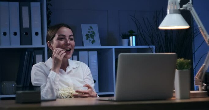 Businesswoman having fun eating popcorn and watching movie on office laptop at night