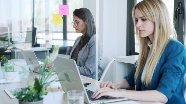 Video of two business women work with laptops on the partitioned desk in the coworking space. Concept of social distancing.