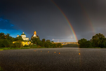 A rare event. Double rainbow in the blue sky over the city. Landscape on the river during the rain. Bridge and beautiful churches. Raindrops on the river. Green trees and grass. 