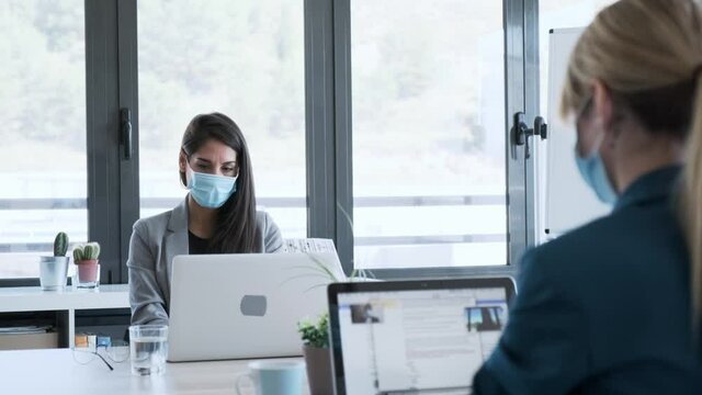 Video of two business women wearing a hygienic face mask while work with laptops in the coworking space. Social distancing concept.