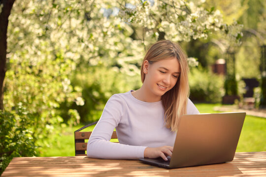 A cute young teen girl is learning online on a laptop while sitting at a wooden table in the back garden of a house.