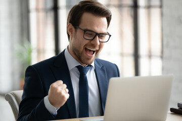 Overjoyed young Caucasian businessman in glasses look at laptop screen triumph read good news email, excited male CEO feel euphoric get amazing unexpected business offer working on computer in office