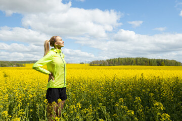 Fototapeta premium A girl in a bright sports jacket rests after training against the background of a blooming rapeseed field