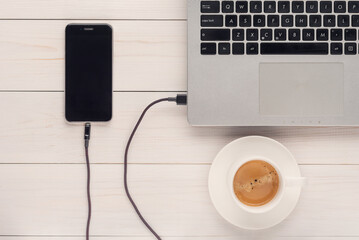 cup of coffee, mobile phone is charging from a computer on a white wooden surface