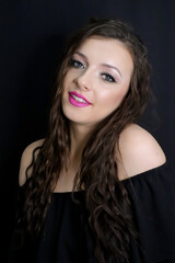 Naklejka premium beautiful brunette young girl happily smiling, looking at camera with fresh make up on black background, pink lipstick curly hair over shoulders, in black dress studio shot, vertical image