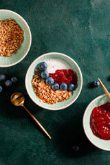 Top view of breakfast with granola, yoghurt, strawberry jam, chia seeds and blueberry on a dark green table.