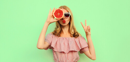 Summer portrait of young woman showing juicy grapefruit blowing red lips sending sweet air kiss over green background