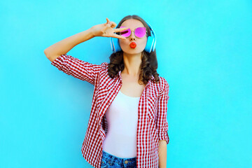 Portrait of attractive young woman in wireless headphones listening to music blowing red lips sending sweet air kiss wearing a pink sunglasses on colorful blue background