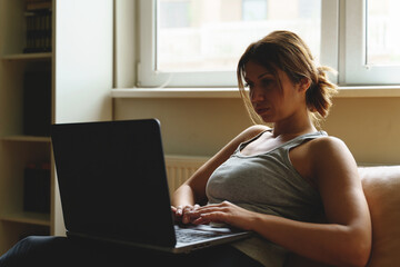 Young adult caucasian woman working on the laptop computer from home sitting on the sofa bed in day taping online female freelancer or shopping online efficiency and expertise making money concept