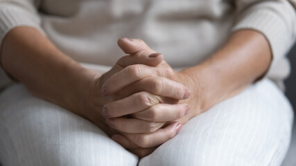 Close up of senior woman sit on couch at retirement house hold hands joined feeling lonely or abandoned, mature 60s female believer praying at home, thinking pondering, elderly solitude concept