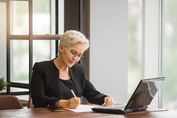 beautiful stylish woman in age in a black suit sits at work with a laptop