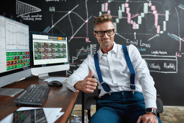Passionate to perform. Middle-aged caucasian trader in glasses looking at camera, showing OK sign while sitting by desk in front of computer. Blackboard full of charts in background.