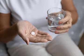 Close up of unhealthy elderly woman hold pill and glass of water feeling unwell sick at home, ill...