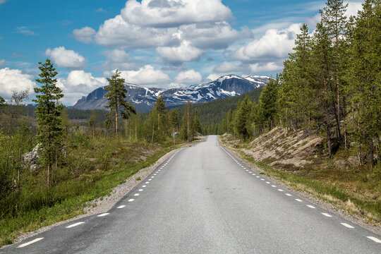 BD 805, a narrow, lonely road leading through the arctic wilderness of Lapland, Sweden, connecting Jokkmokk and Kvikkjokk. A rugged mountain range covered with snow is visible in the background.