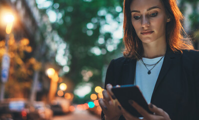 Confident young woman in black suit working documents using smartphone on background lights evening...