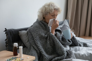 Sick older woman sit in bed blow running nose, suffer from flu or cold at home, ill mature female...