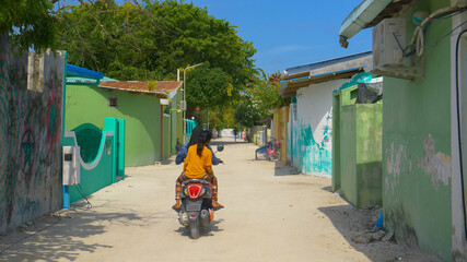 Two locals ride an old scooter down an empty road in a poor village in Maldives