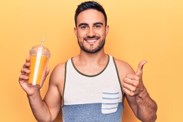 Fototapeta premium Handsome man with beard wearing sleeveless t-shirt drinking glass of healthy orange juice smiling happy and positive, thumb up doing excellent and approval sign