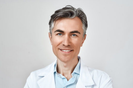 Portrait of happy seniour professional caucasian doctor in medical uniform looking at camera and smiling while standing against grey background, posing in studio