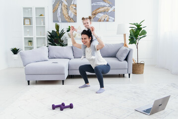 Beautiful mommy and charming little daughter are smiling while doing fitness exercises together at home.