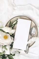 Festive summer wedding scene. Marble table setting with cutlery, olive branches, white peony flowers, stoneware plate and silk ribbon. Blank restaurant menu card mockup. Flat lay, top view, vertical.