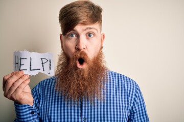 Redhead Irish man with beard holding paper with help message over blue background scared in shock...