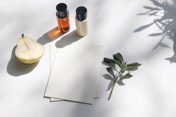Summer stationery still life scene. Cut pear fruit, cosmetic oil, cream bottles and olive tree branch. White table. Blank paper cards, invitations mockup scene. Long shadows overlay. Flat lay, top