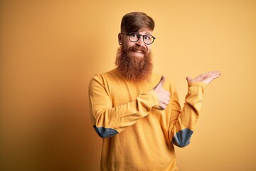 Handsome Irish redhead man with beard wearing glasses over yellow isolated background Showing palm hand and doing ok gesture with thumbs up, smiling happy and cheerful