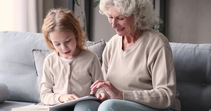 Cute little redhead child girl learning reading paper book with happy middle aged grandmother, sitting together on couch indoors. Smiling senior old woman feeling proud of smart small granddaughter.