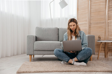 Focused young freelance woman sitting on the table at home with laptop and headphones, working remotely online from home. Concentrated teenage girl studying distantly alone in living room.