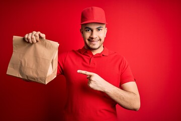 Young handsome delivery man holding paper bag with takeaway food over red background very happy pointing with hand and finger