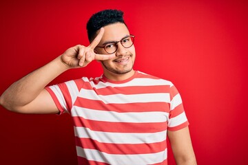 Young handsome man wearing casual striped t-shirt and glasses over isolated red background Doing peace symbol with fingers over face, smiling cheerful showing victory