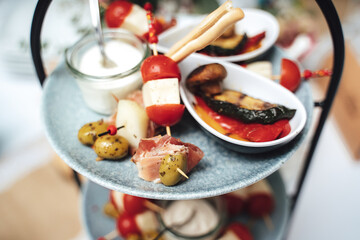 Multi-tiered plate stand with small snacks and ingredients. Stuffed cherry tomatoes, olives and glass of white dip. Celebration, party, wedding