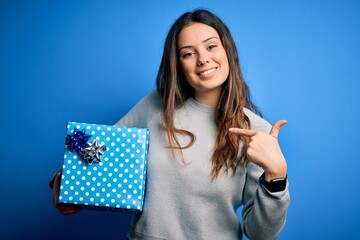 Young beautiful brunette woman holding birthday gift over isolated blue background with surprise...