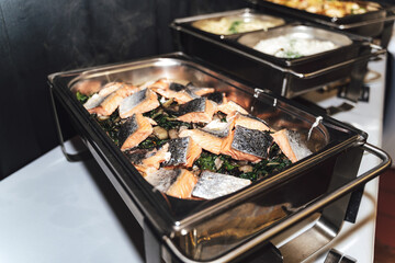 Row of stainless hotel pans on food warmers with various meals. Smoked pieces of salmon fish....