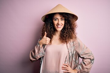 Young beautiful woman with curly hair and piercing wearing traditional asian conical hat doing happy thumbs up gesture with hand. Approving expression looking at the camera showing success.
